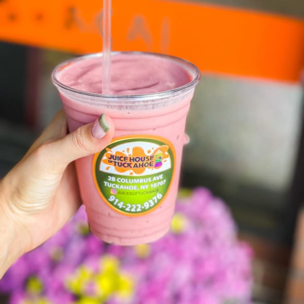 Hand holding a pink smoothie from Juice House of Tuckahoe with a straw, against a blurred floral background.