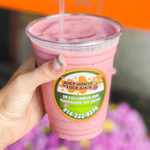 Hand holding a pink smoothie from Juice House of Tuckahoe with a straw, against an orange background.