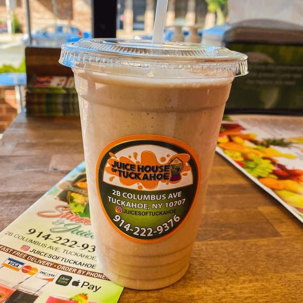 A Juice House of Tuckahoe smoothie in a plastic cup with a straw, on a wooden table.